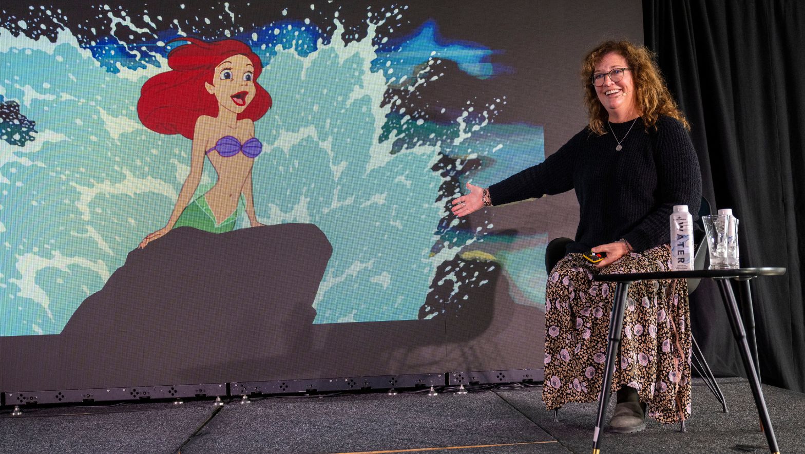 A seated woman gestures to a screen behind her which depicts an animated mermaid character