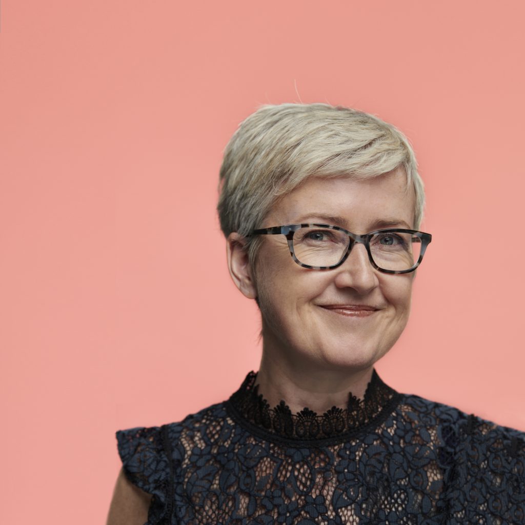 A smiling person with short, light hair wears glasses and a dark, lacy top against a soft pink background, conveying a cheerful, approachable mood.