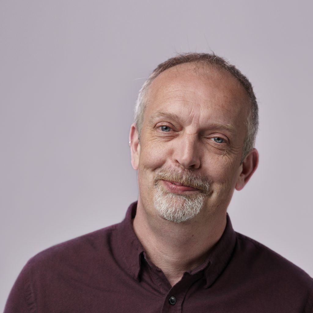 A man with a goatee and short hair looks at the camera with a slight smile. He's wearing a maroon shirt against a plain background.
