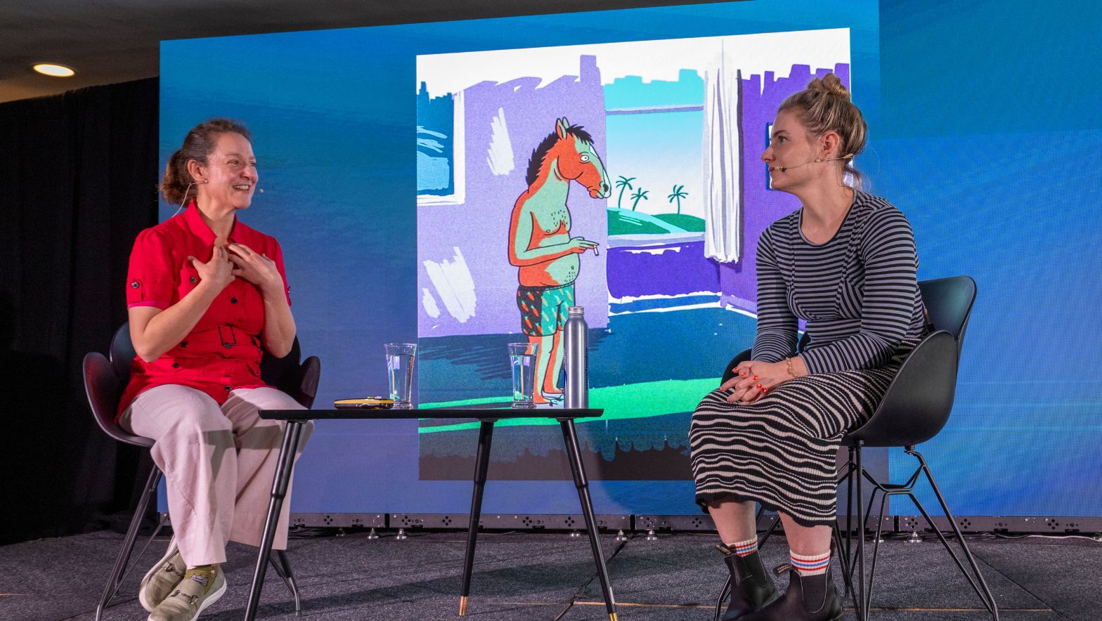 Two individuals wearing headset microphones sit and chat on stage in front of an illustration of the animated television series 