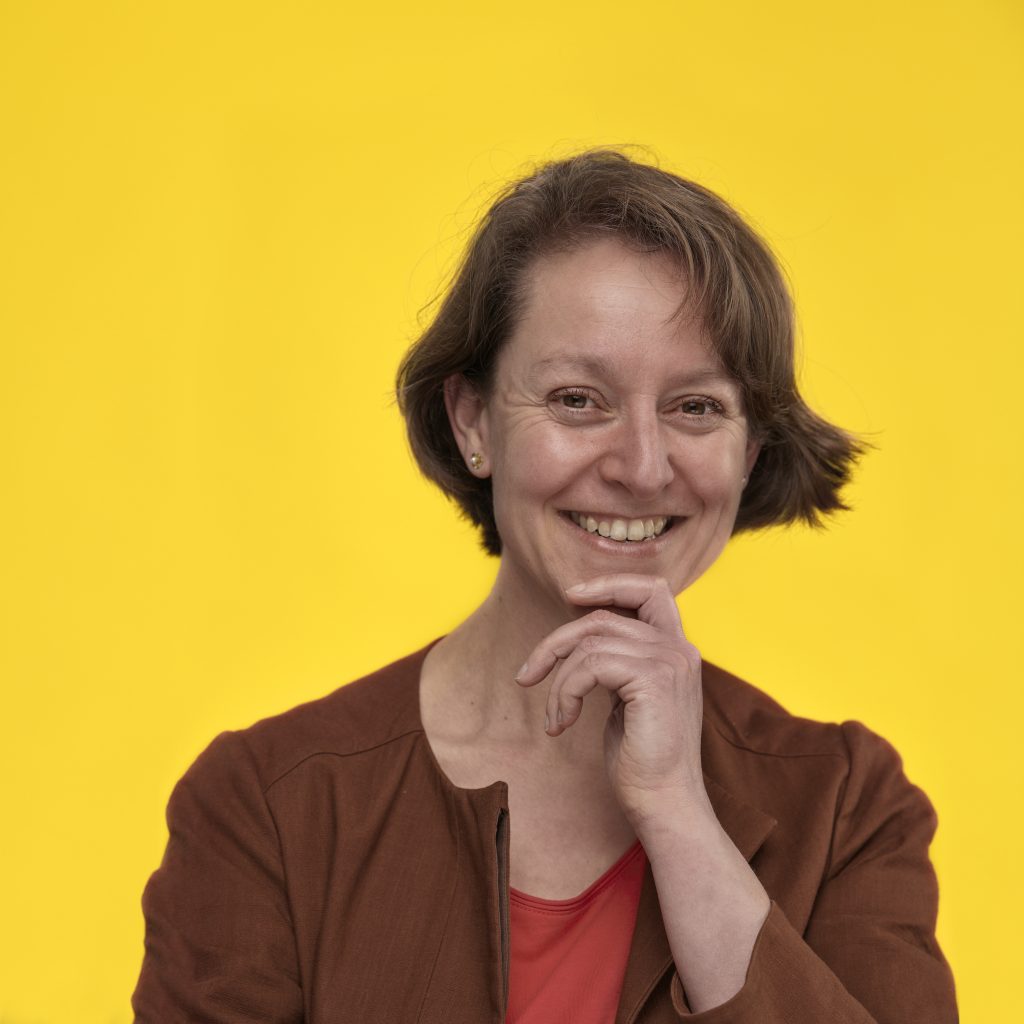 Smiling person with short brown hair and hand on chin, wearing a brown jacket and red top, stands against a bright yellow background.