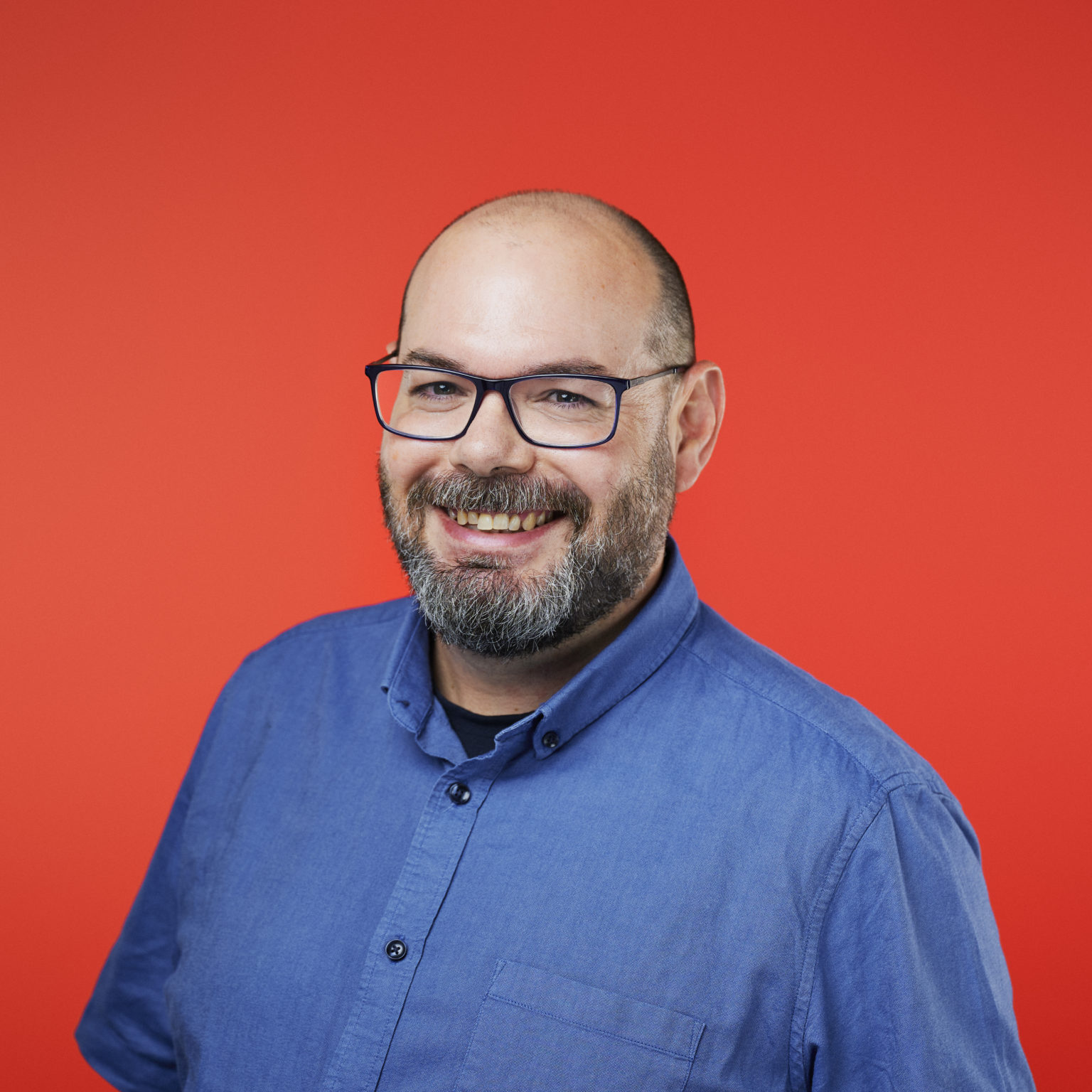 A smiling man with glasses and a beard against a bright red background. He wears a blue button-up shirt, creating a friendly and approachable tone.