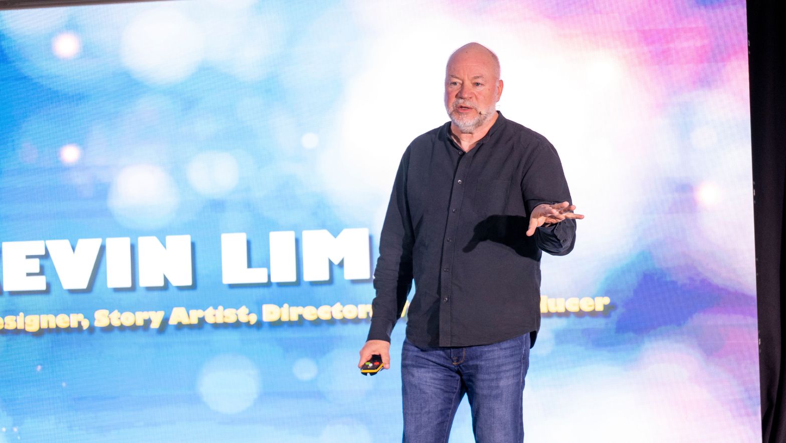 A man stands in front of a large screen wearing a headset microphone. 