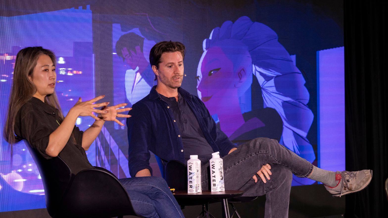 Two individuals wearing headset microphones are seated in front of a large screen with a vivid image from the movie K-Pop Demon Hunters.