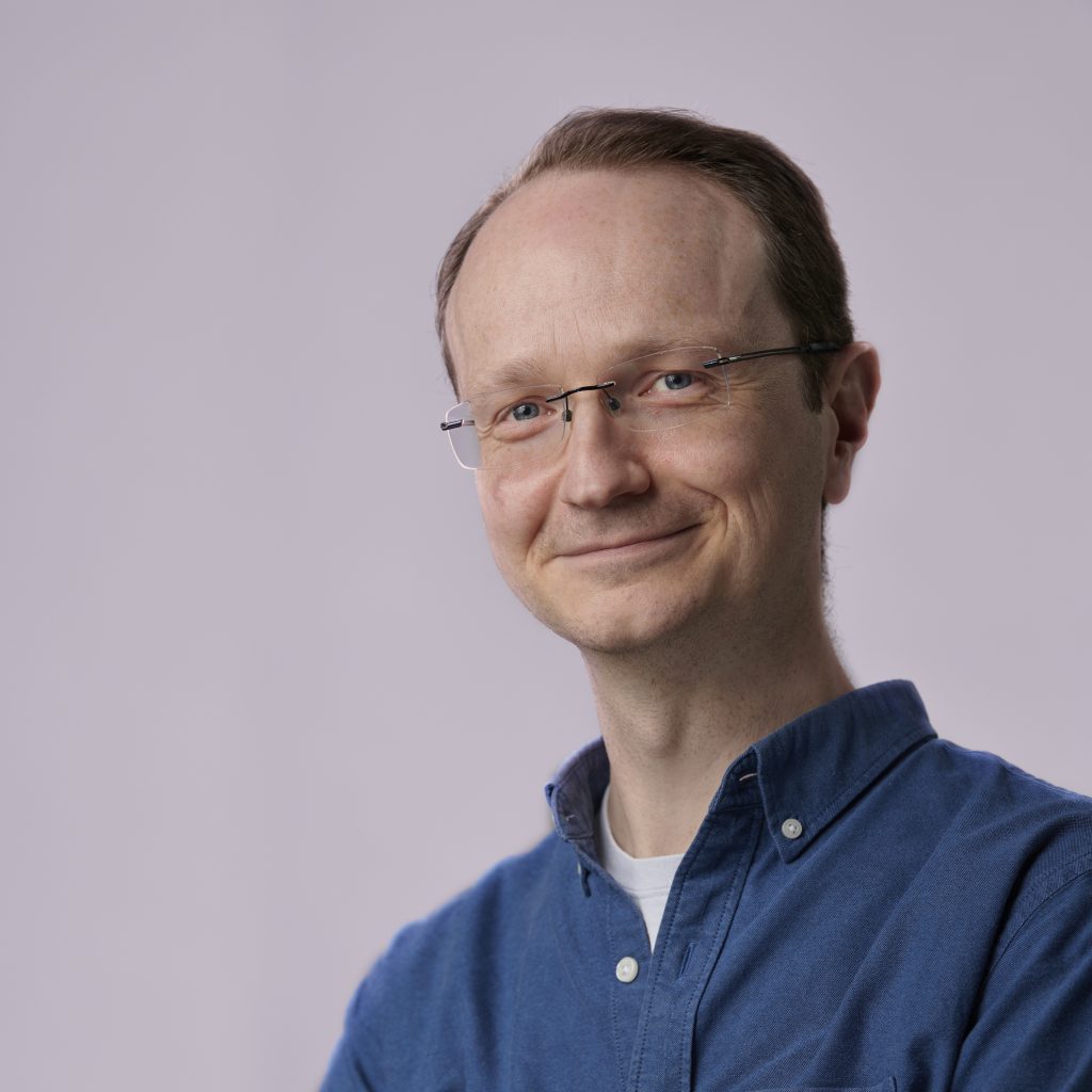 Smiling man with short hair and glasses wearing a blue button-up shirt against a plain light background, conveying a friendly and approachable demeanor.