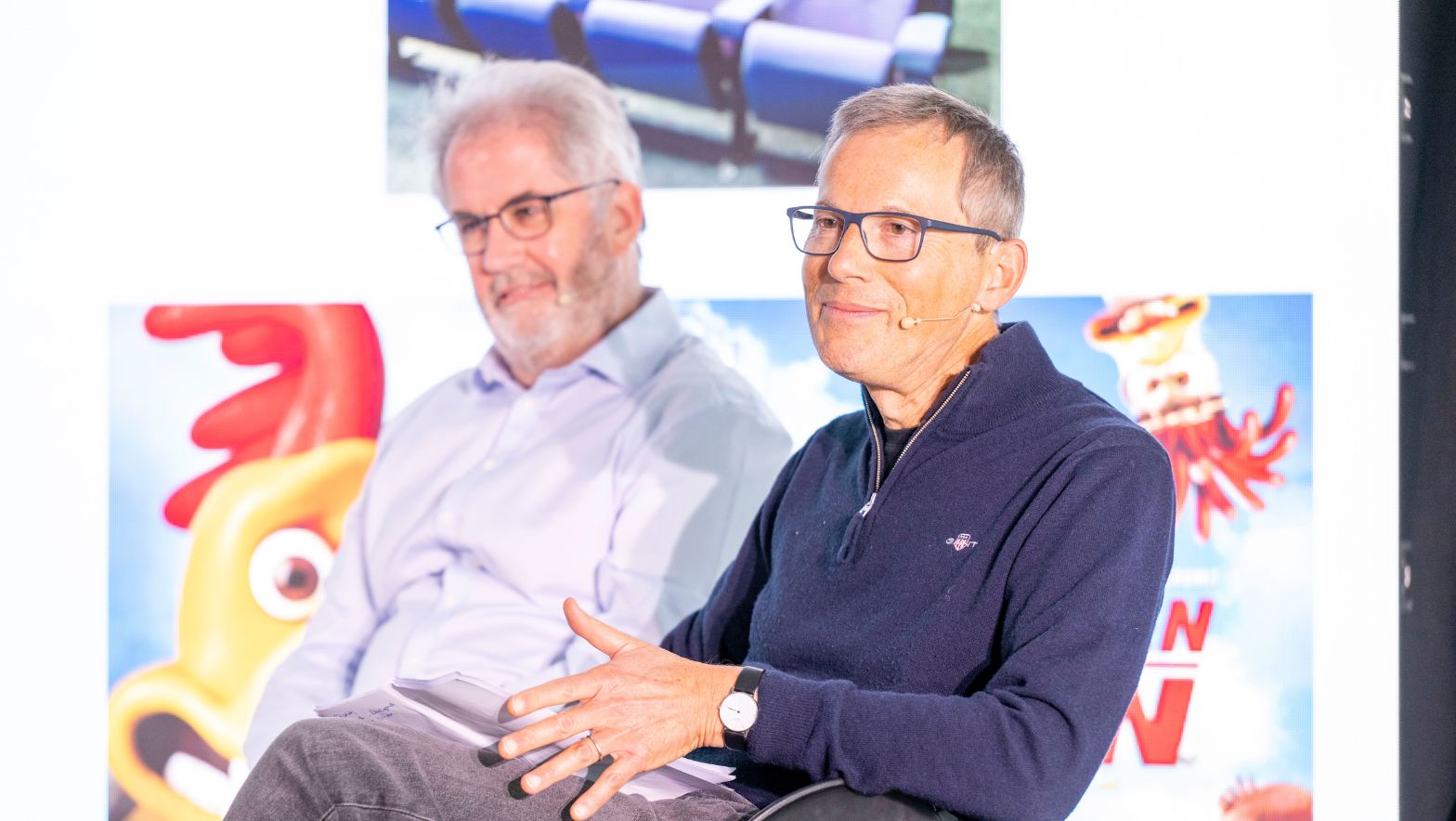 Two individuals in smart attire are seated in what appears to be a corporate setting. Both wear microphones attached to their heads. 