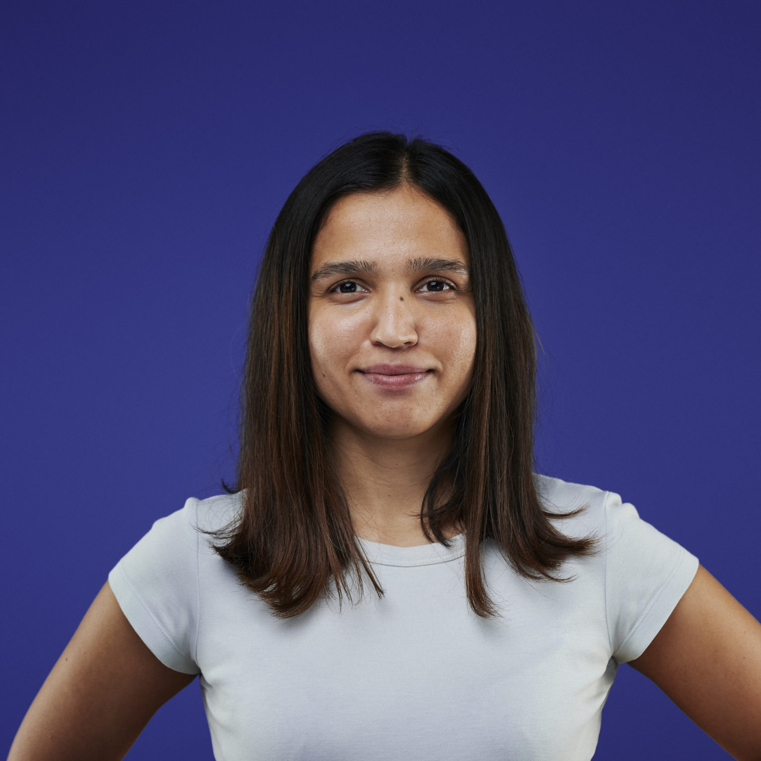A person with long dark hair, wearing a light shirt, smiles subtly against a solid blue background. Their hands are on their hips, conveying confidence.