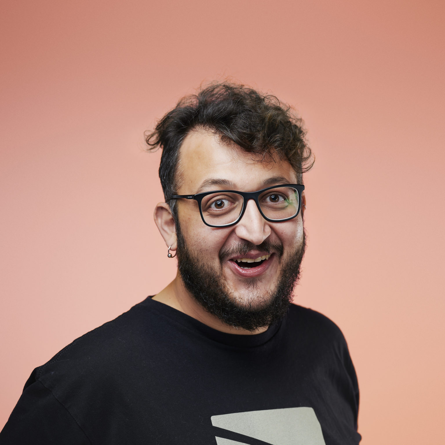 A smiling man with curly hair, glasses, and a beard, wearing a black T-shirt, stands against a peach-colored background, conveying a cheerful mood.