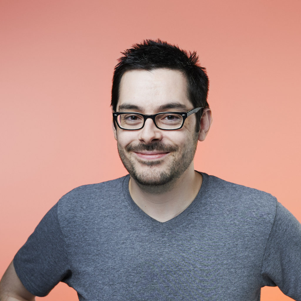 Smiling man with short dark hair and glasses in a gray t-shirt stands against a peach background, exuding a cheerful, approachable vibe.