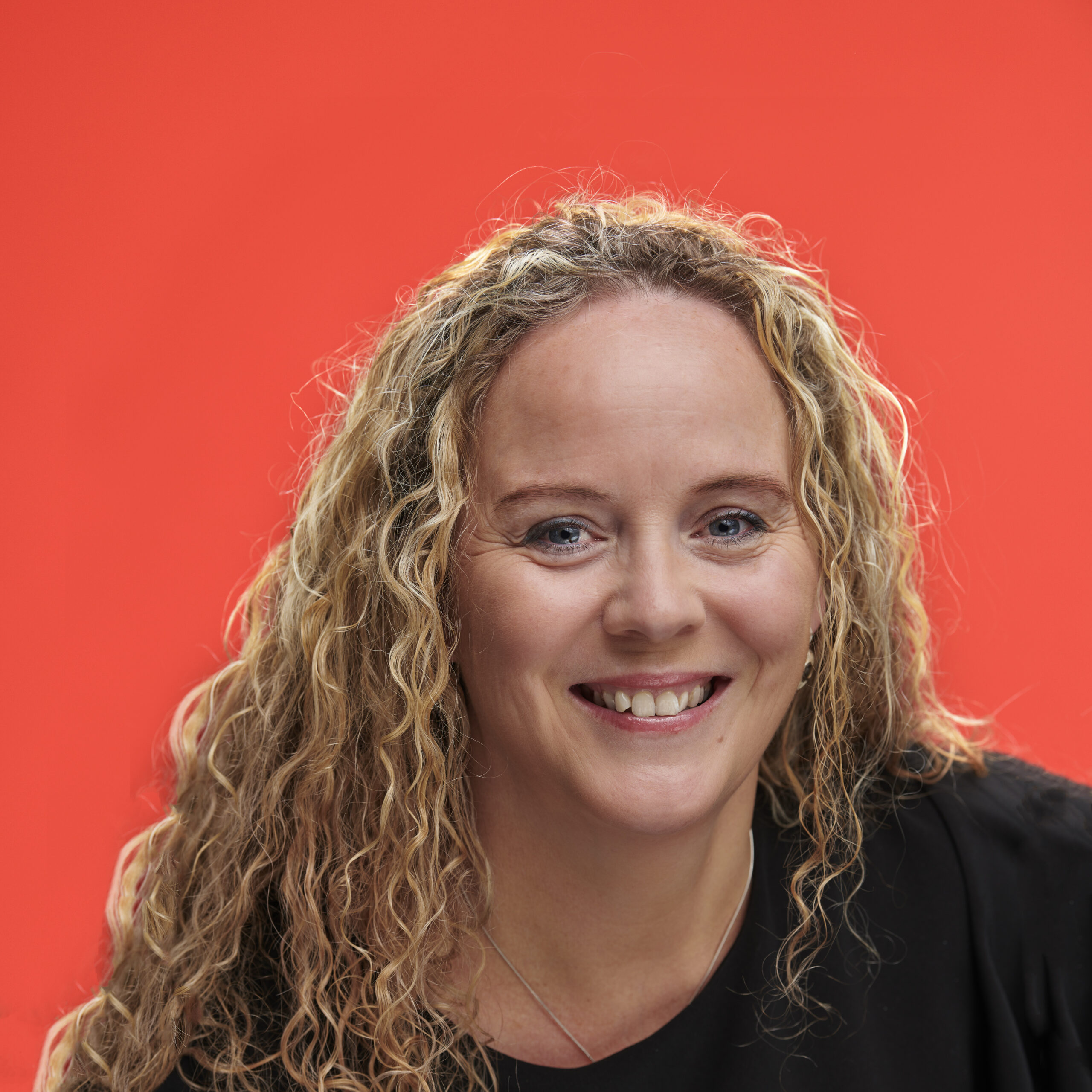 Smiling woman with curly blonde hair against a vibrant orange background. She wears a black top, conveying a cheerful and welcoming tone.
