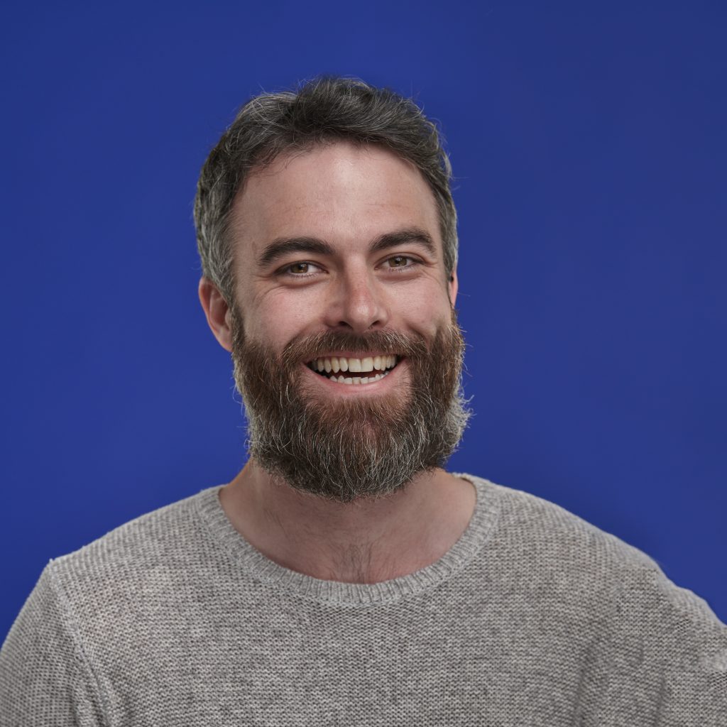A man with a beard and gray sweater smiles warmly against a vibrant blue background. His expression conveys happiness and friendliness.
