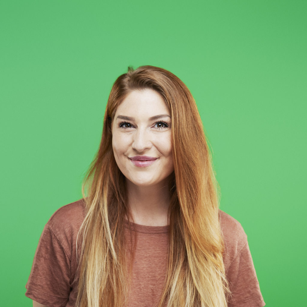 Smiling woman with long, red hair wearing a brown shirt, stands against a bright green background, conveying a cheerful and vibrant mood.
