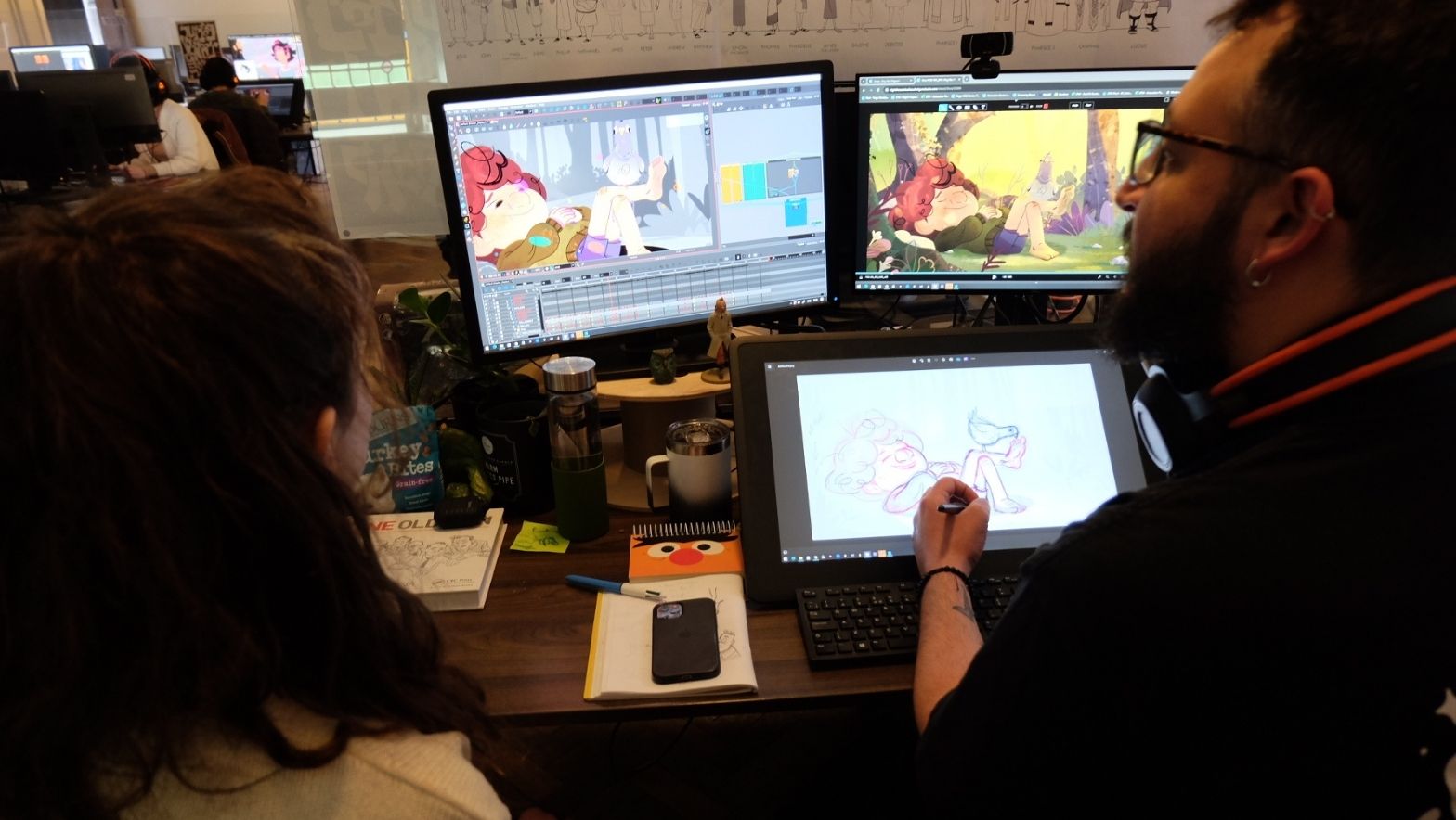 Two individuals face three computer screens, all with a vivid illustration of a young boy and bird. 