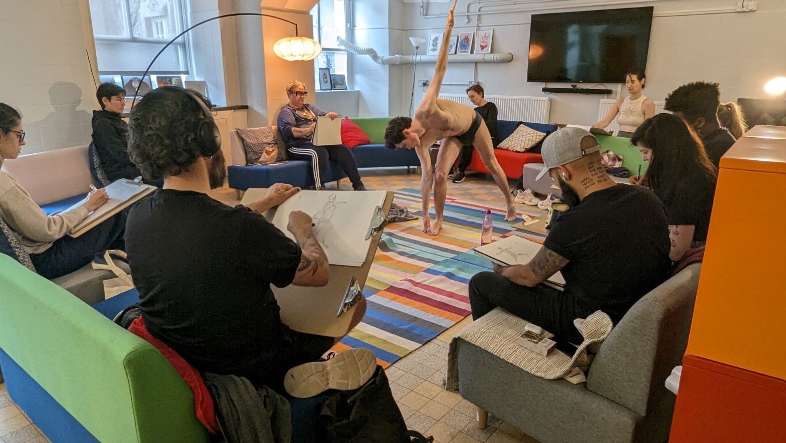 A group of people are seated and sketching a model in a stretched pose in a cosy setting.