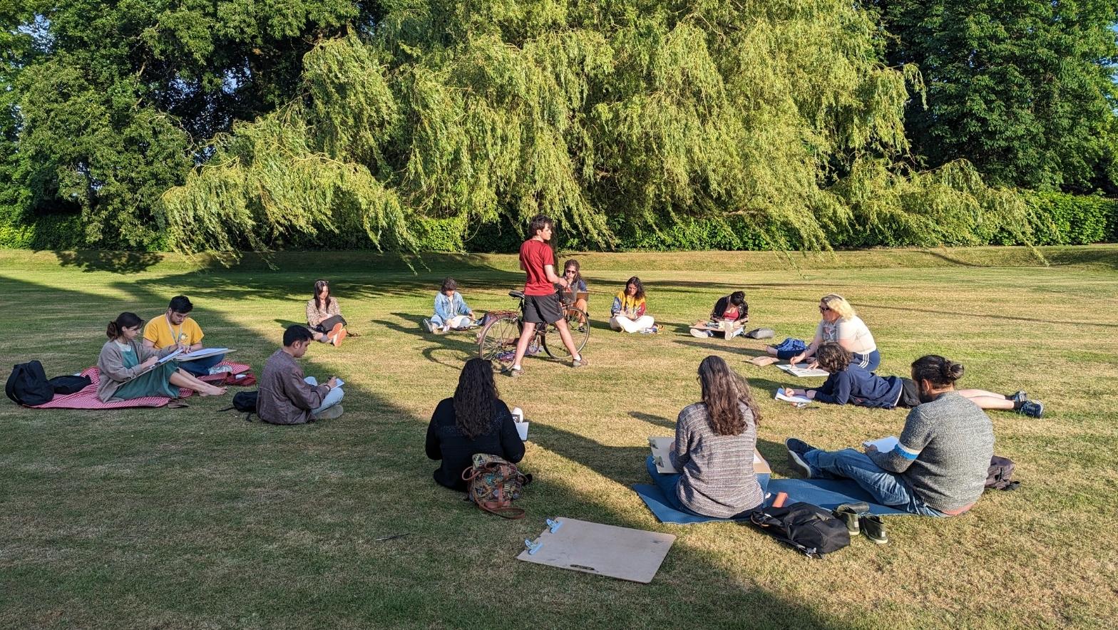 A group of people on a sunny lawn are seated in a circle, sketching a person posing with a bicycle. 