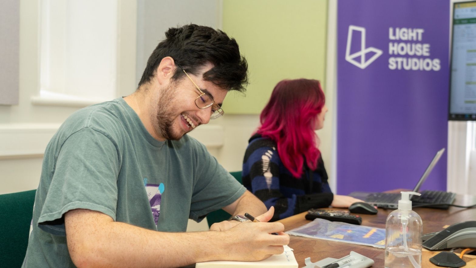 Two people are seated at a large table. One is smiling as he sketches. 