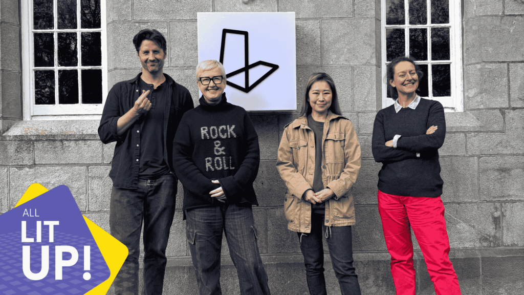 Four smiling individuals pose in front of a lightbox that displays the logo for Lighthouse Studios. In the corner is a graphic logo containing the words 'All Lit Up!' representing the logo for the studio's podcast.