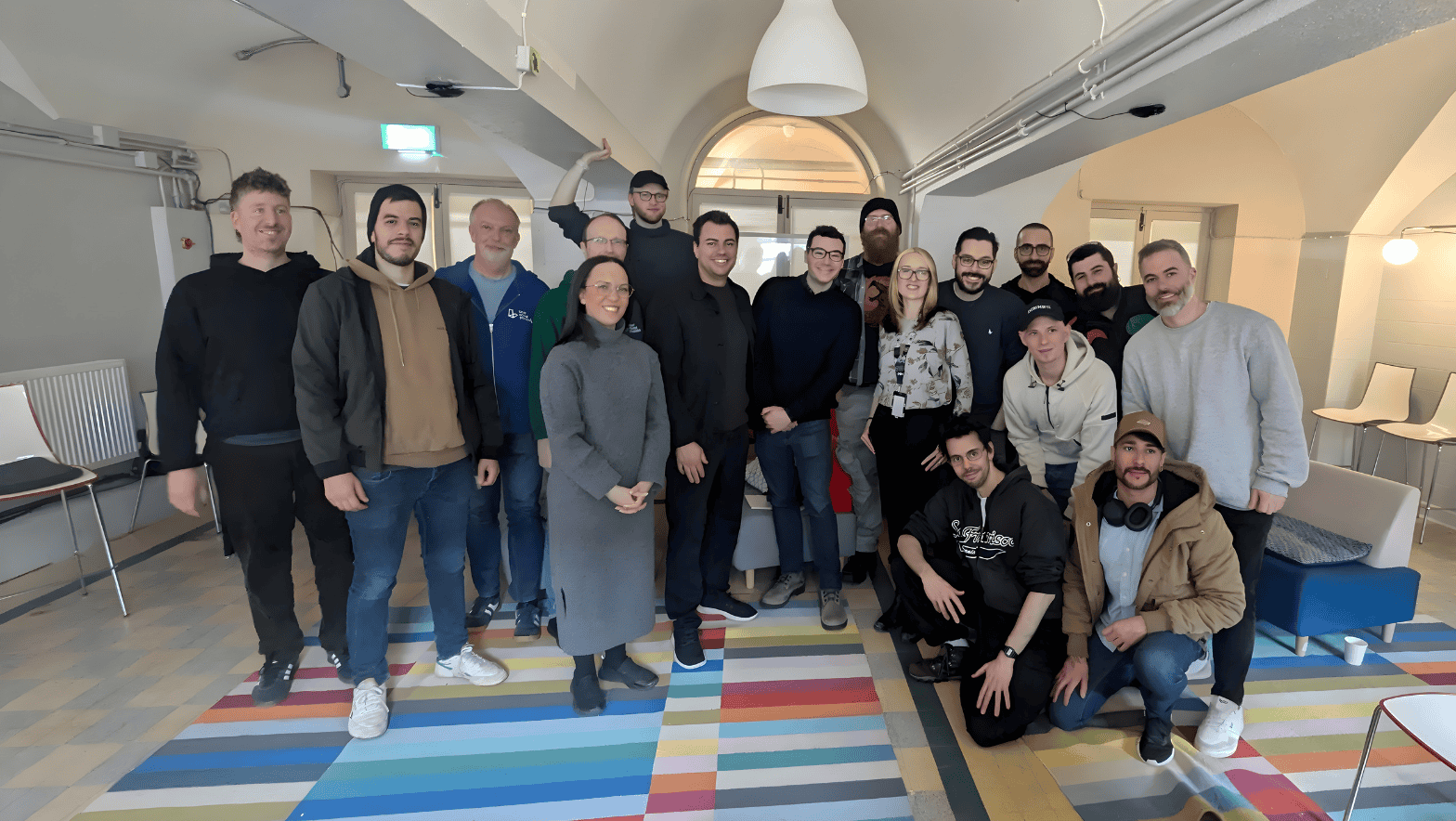 A diverse group of 18 people smiles in a cozy room with arched ceilings. They stand on a multicolored striped rug, conveying a friendly, team spirit.